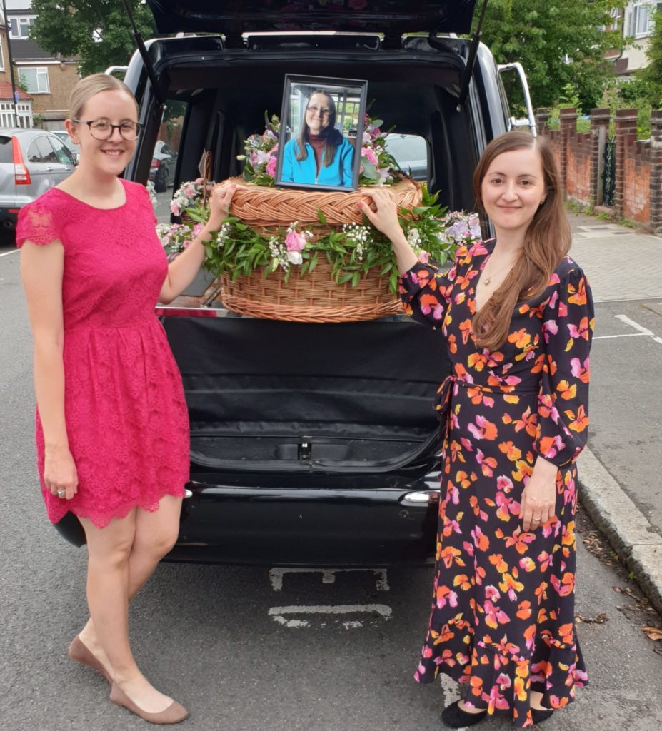 Naomi and her sister stand either side of their mum's coffin inside the hearse. The wicker coffin is fringed with white and pink roses and a photo of their mum smiling is on top of the coffin. They are both smiling and each have one hand on the coffin.